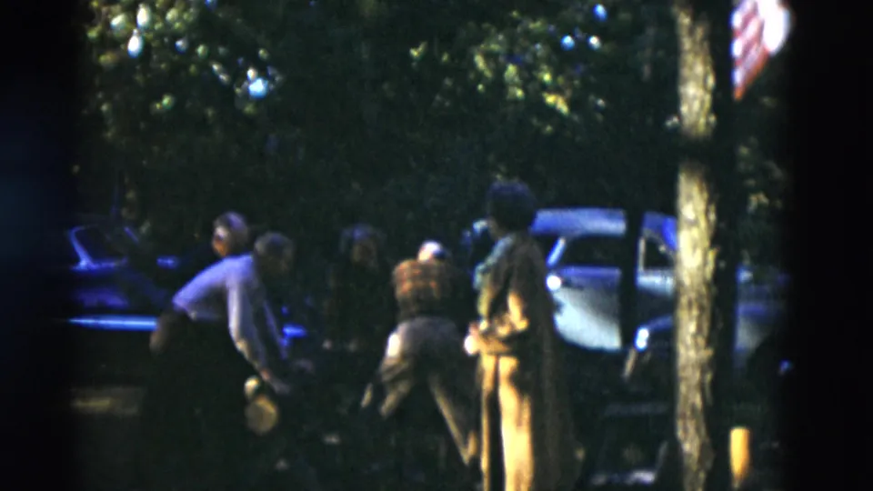 Several older adults gather on a shaded Somerset lawn, with one person bending forward near a car and an American flag partly visible.