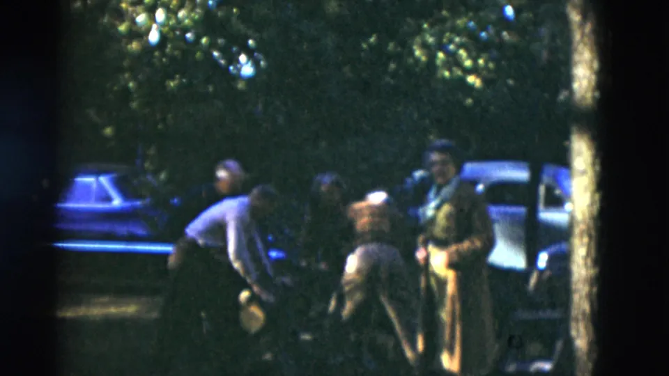 Older adults remain gathered on a wooded lawn in Somerset, Kentucky, with older cars behind them in grainy 8mm footage.