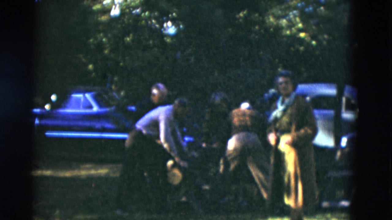 In 1975 Somerset, Kentucky home-movie footage, older adults gather on a shaded lawn near parked cars, with one person bending forward.