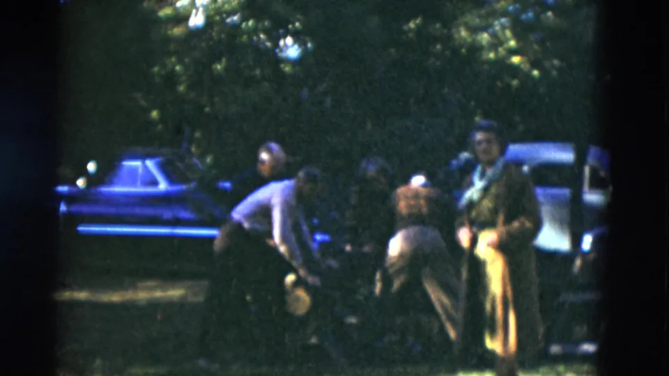 In 1975 Somerset, Kentucky home-movie footage, older adults gather on a shaded lawn near parked cars, with one person bending forward.
