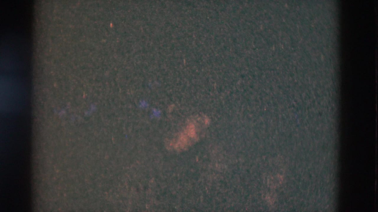 Dark, grainy frame with faint warm patches and vertical film-strip shadows.