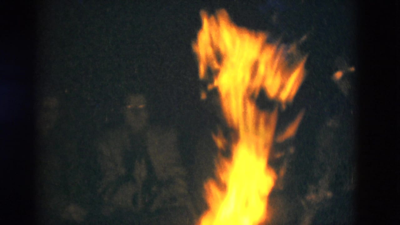 A vertical campfire flame glows against a dark outdoor background, with faint camper silhouettes visible in the 1975 Kentucky home-movie frame.
