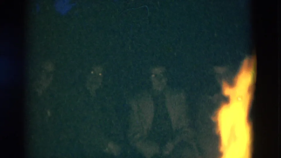 The campfire burns at the edge of the frame while several dimly lit campers sit in the shadowed background of the 8mm night scene.