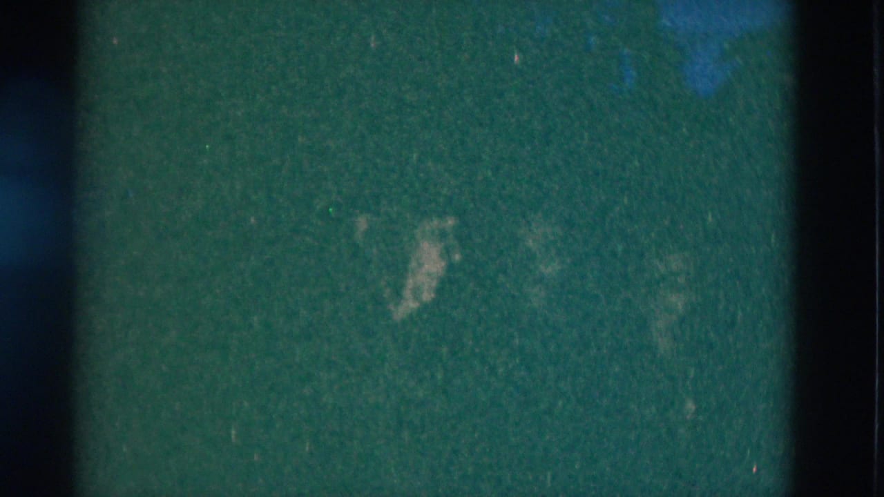 Dark green-tinted frame with faint figures against a shadowed background.