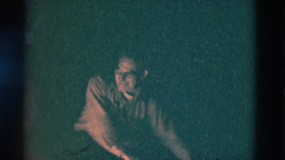 Older man seated outdoors at night in 1975 Somerset, Kentucky footage, his face and jacket dimly lit by firelight.