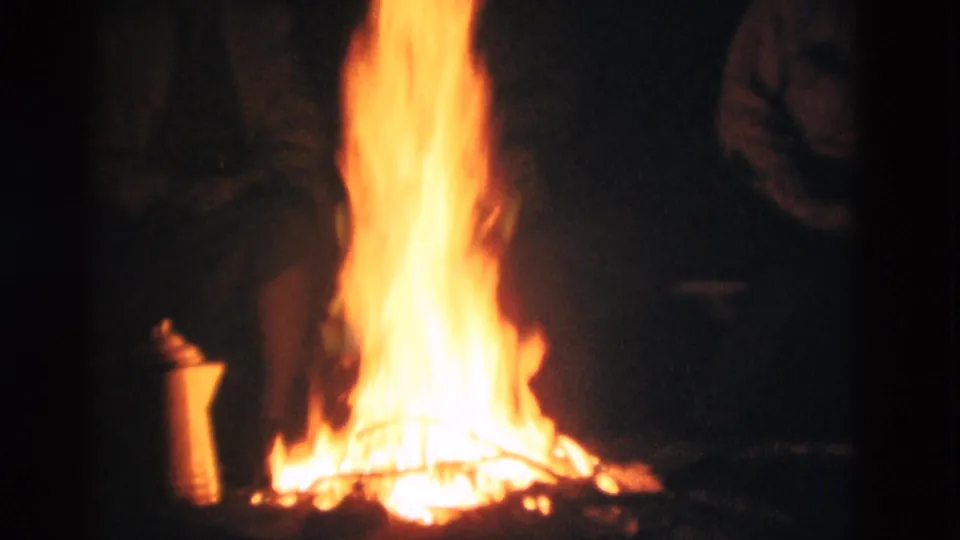 1975 8mm home-movie frame from Somerset, Kentucky, showing a bright campfire at night with adults standing in shadow around it.
