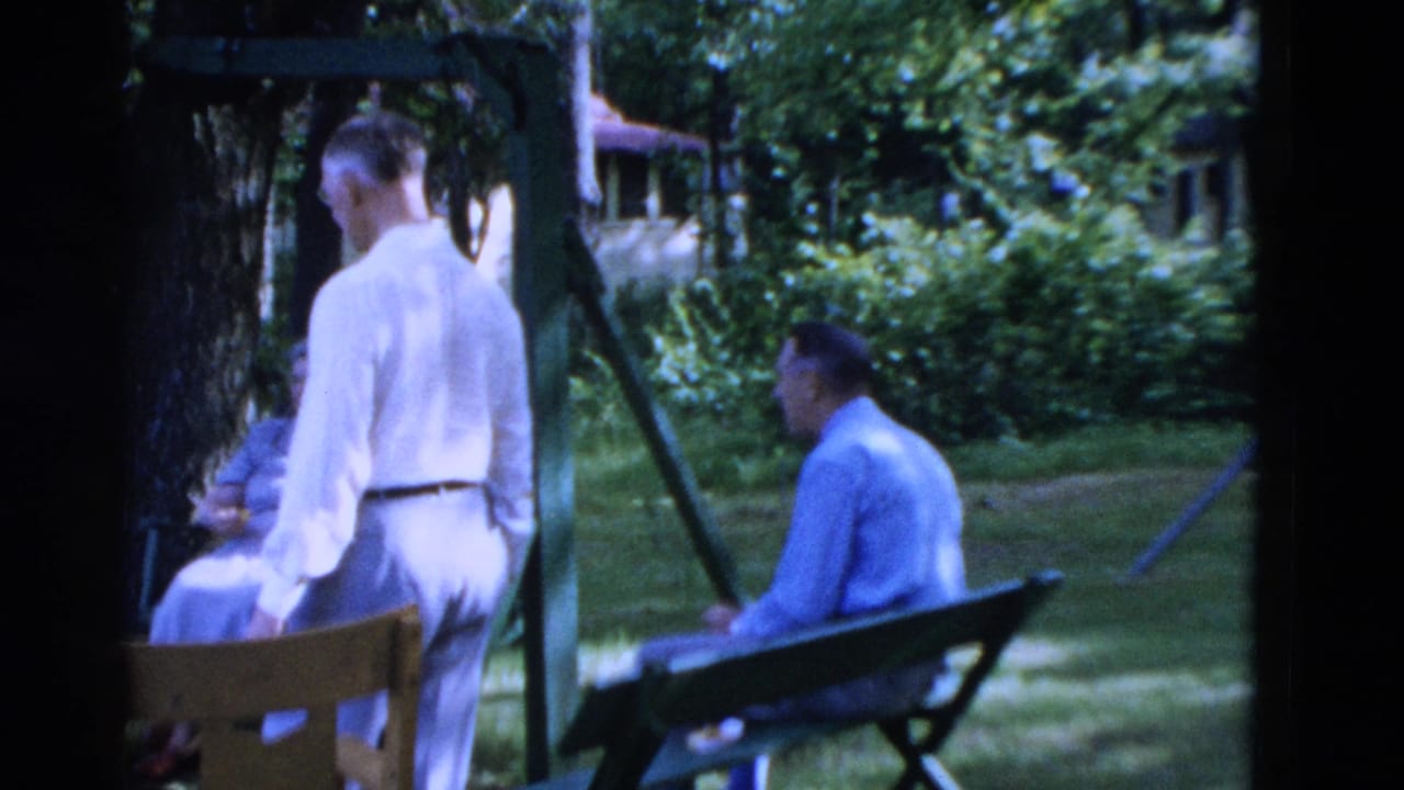 1975 Somerset, Kentucky home-movie frame showing adults in light clothing gathered in a shaded backyard with a bench and swing frame.