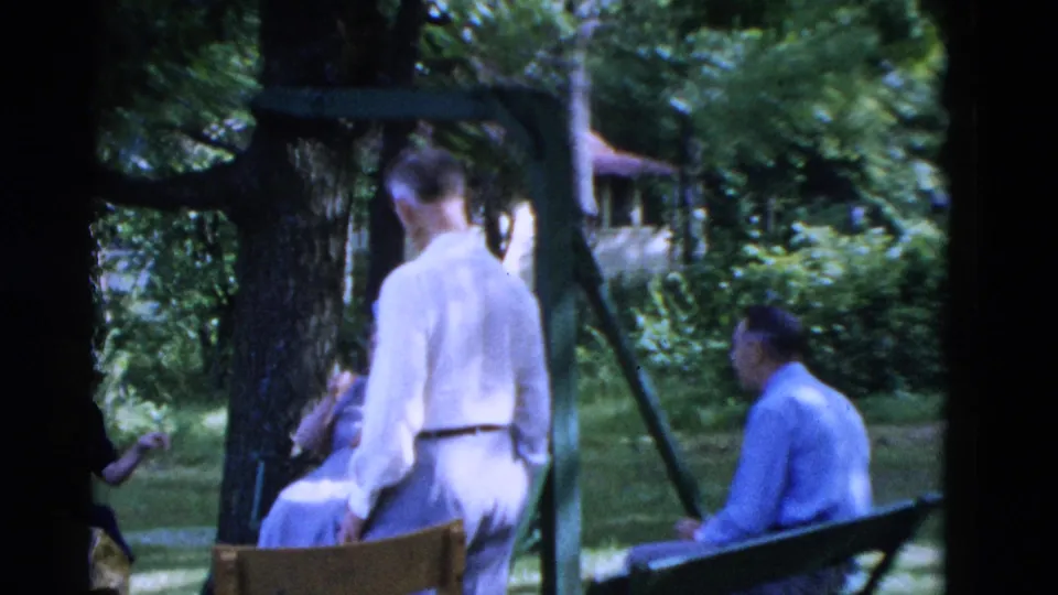 Soft 8mm frame from the Somerset, Kentucky gathering, showing adults conversing in a shaded yard beside a bench and swing support.