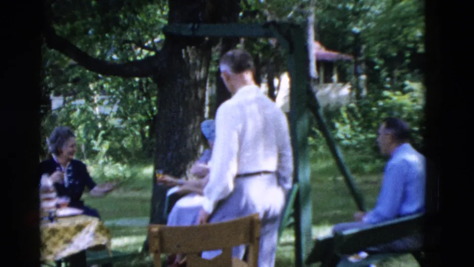 1975 8mm home-movie frame from Somerset, Kentucky, showing adults seated and talking in a tree-shaded backyard picnic setting.