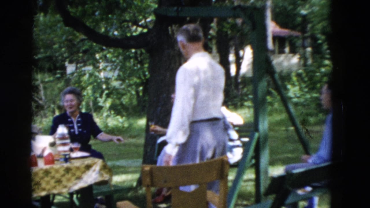 Adults gathered at a picnic table under trees while a man stands nearby.
