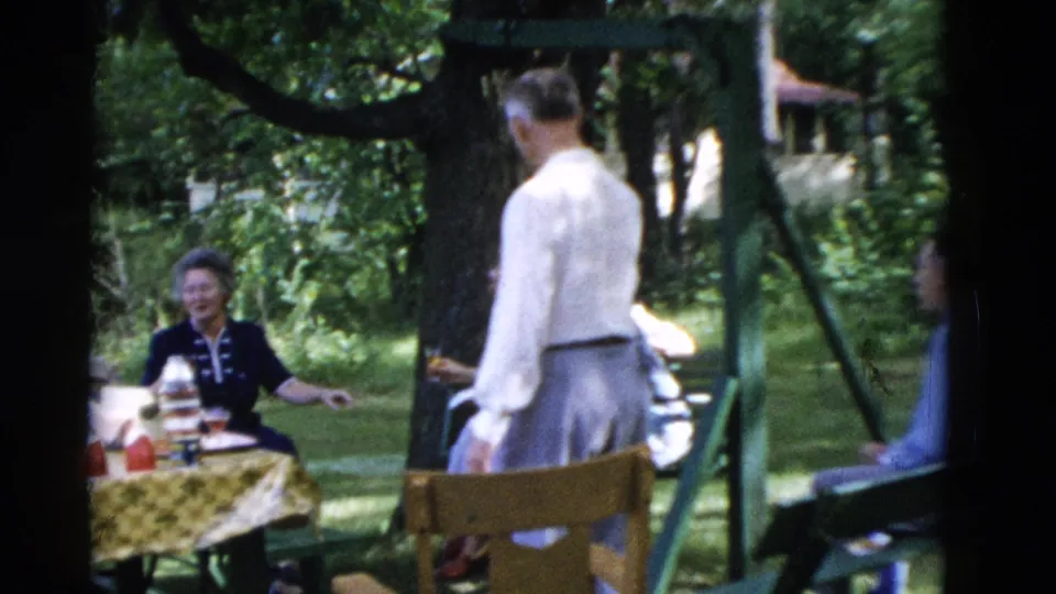 1975 Somerset, Kentucky home-movie frame showing adults around a backyard picnic table with bottles and cups under trees.