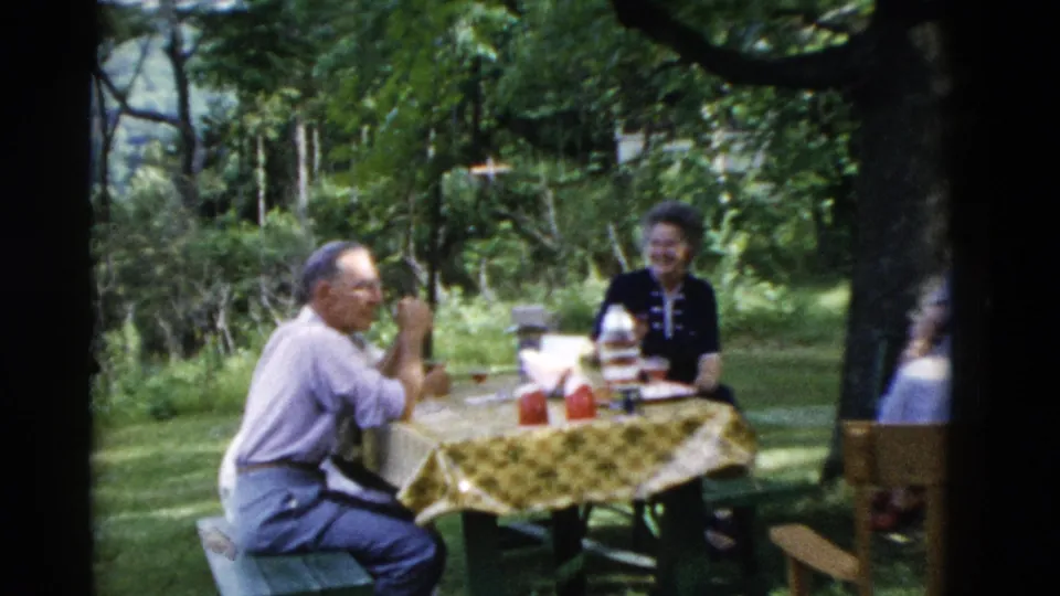 1975 Somerset, Kentucky 8mm frame showing older adults around a picnic table with a yellow patterned cloth in a wooded yard.