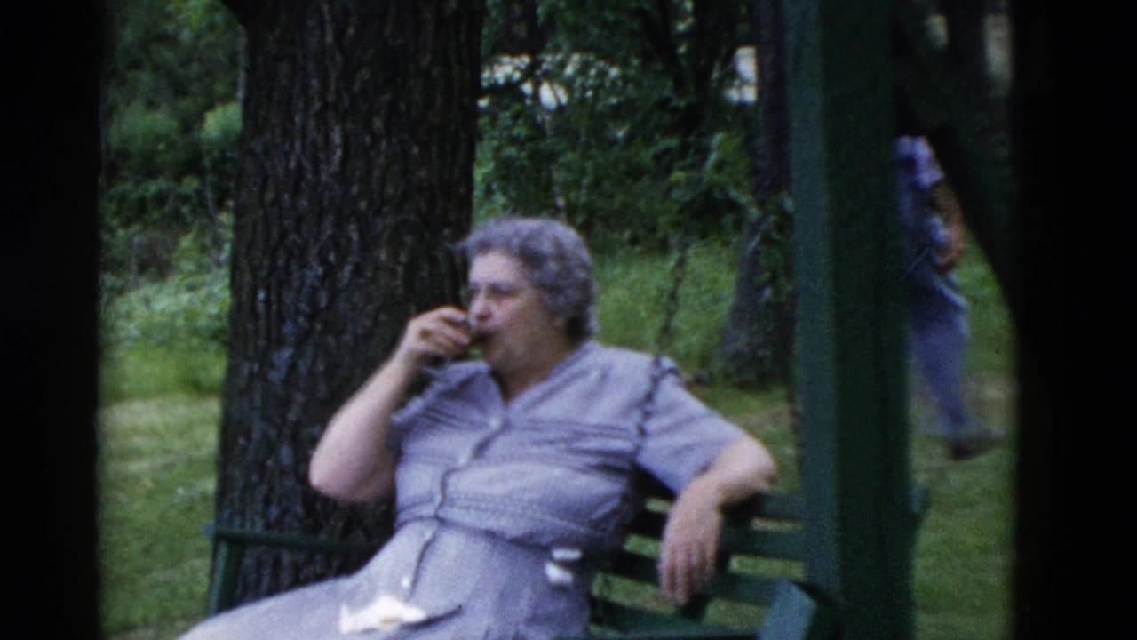 Older woman in a light dress sits on a green porch swing in a wooded yard, drinking from a small glass.