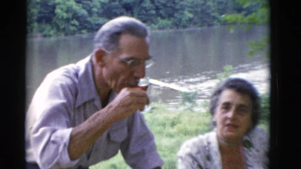 1975 home-movie frame of an older man drinking near a seated woman, with a wooded waterbank in the background.