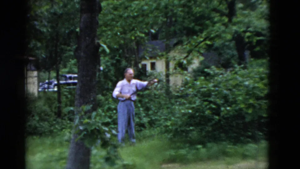 Older man stands in a green yard among trees and shrubs, with a small building partly visible in the background.