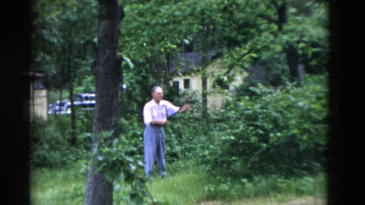 In Somerset, Kentucky home-movie footage from 1975, a man stands among shrubs in a wooded yard with cars and a small building behind him.