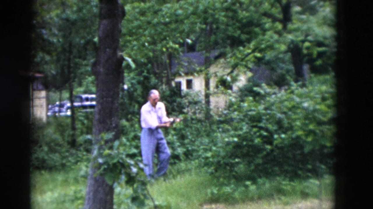 A man in a white shirt and blue pants appears to tend or examine plants in a wooded Somerset, Kentucky yard in 1975 footage.