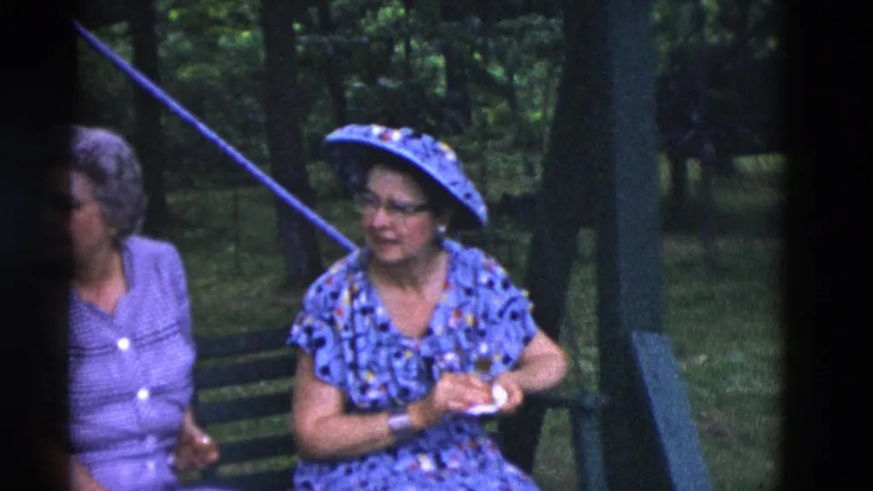 Two women sit on green benches, one wearing a patterned blue dress and matching hat, in 1975 Somerset, Kentucky home-movie footage.