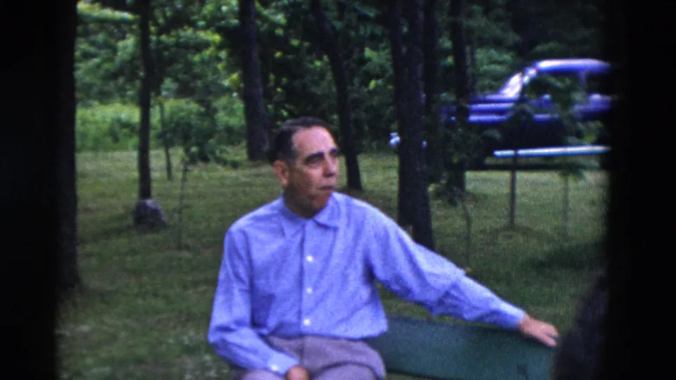 A man in a light blue shirt sits on a green bench in a wooded Somerset, Kentucky yard, with an older car visible in the background.