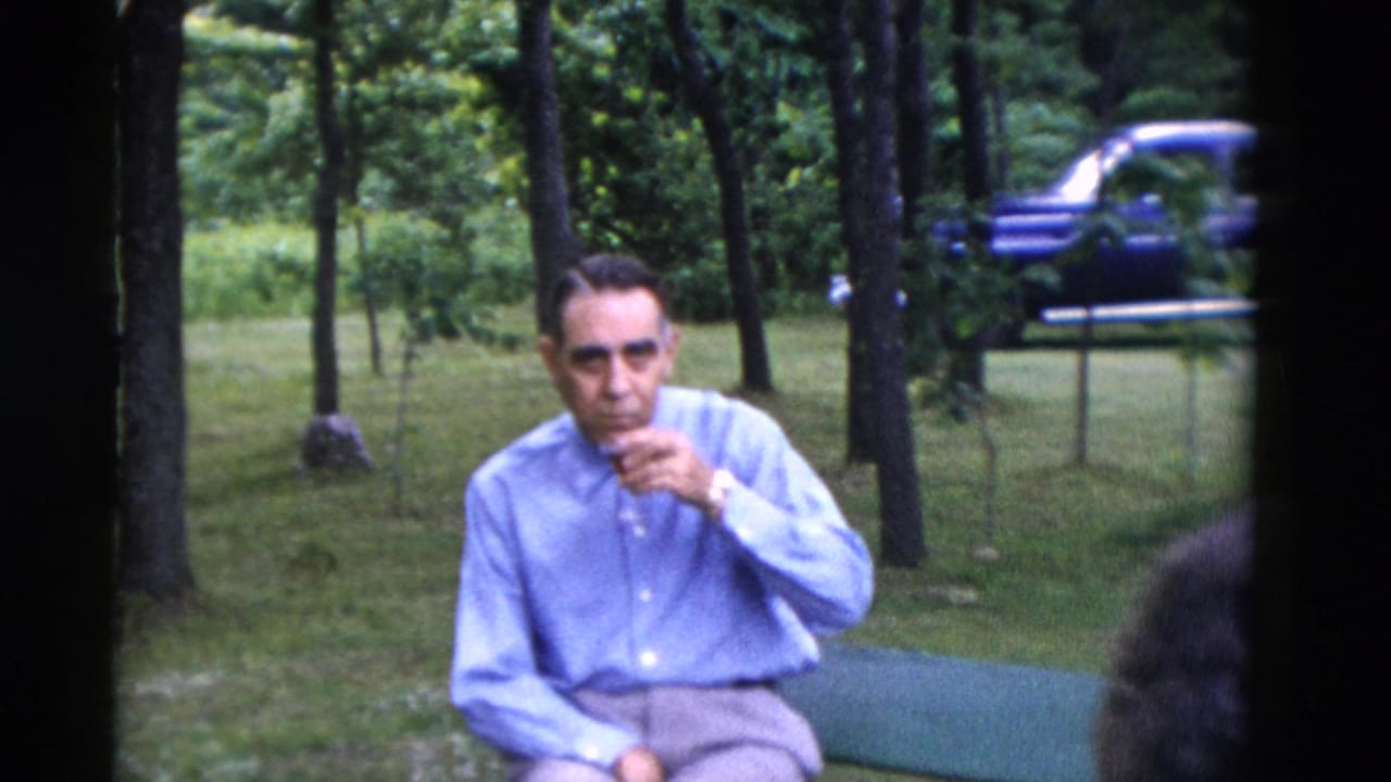 1975 Somerset, Kentucky home-movie frame of a seated man in a light blue shirt sipping a drink near trees, with a blue car behind him.