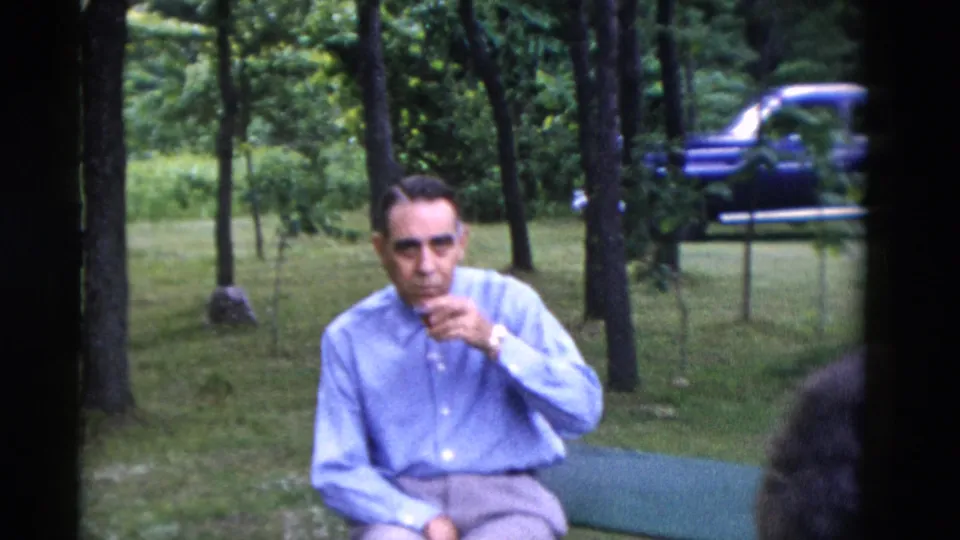 1975 Somerset, Kentucky home-movie frame of a seated man in a light blue shirt sipping a drink near trees, with a blue car behind him.