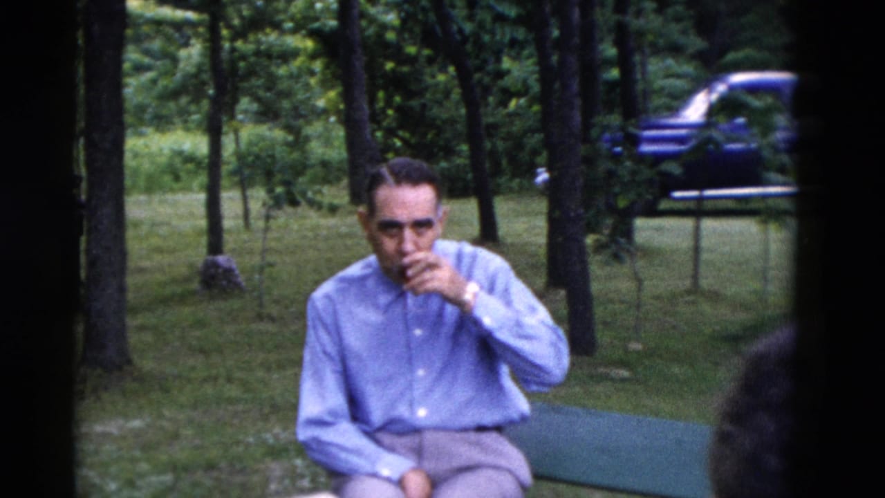 1975 Somerset, Kentucky frame showing a man seated outdoors with a glass at his mouth, trees and a blue car in the background.