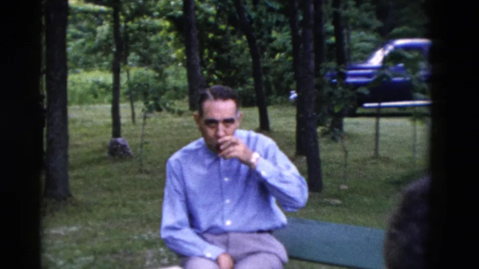 1975 Somerset, Kentucky frame showing a man seated outdoors with a glass at his mouth, trees and a blue car in the background.