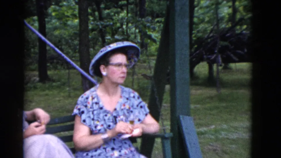 1975 Somerset, Kentucky home-movie frame of a woman in a patterned dress and wide-brimmed hat seated on a green bench in the woods.