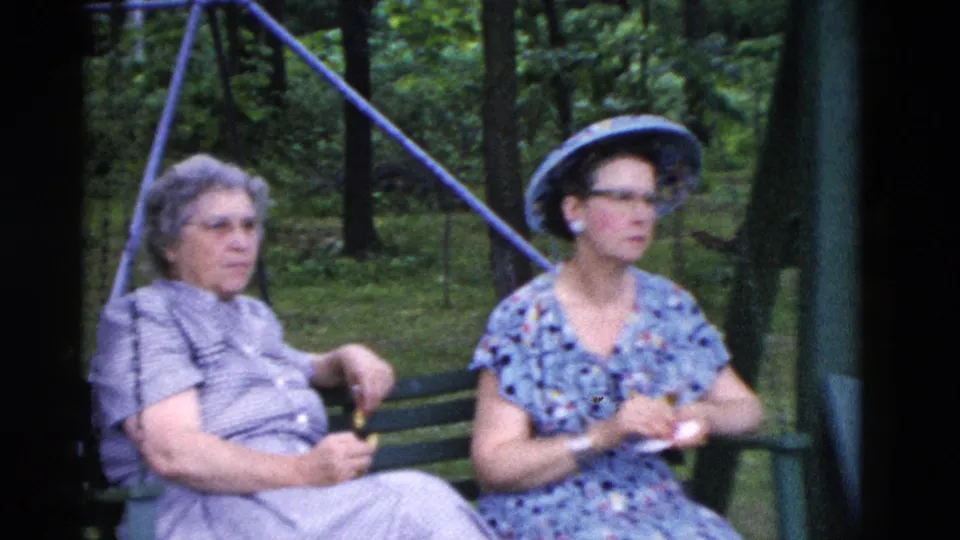 1975 Somerset, Kentucky frame of two women seated on a green bench in the woods, one in a patterned dress and wide-brimmed hat.