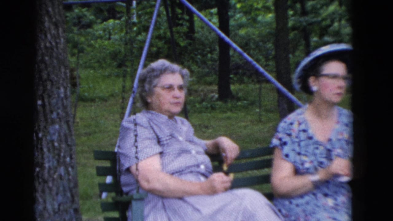 Two women sit on a green park bench between trees, one wearing glasses and a dark hat.