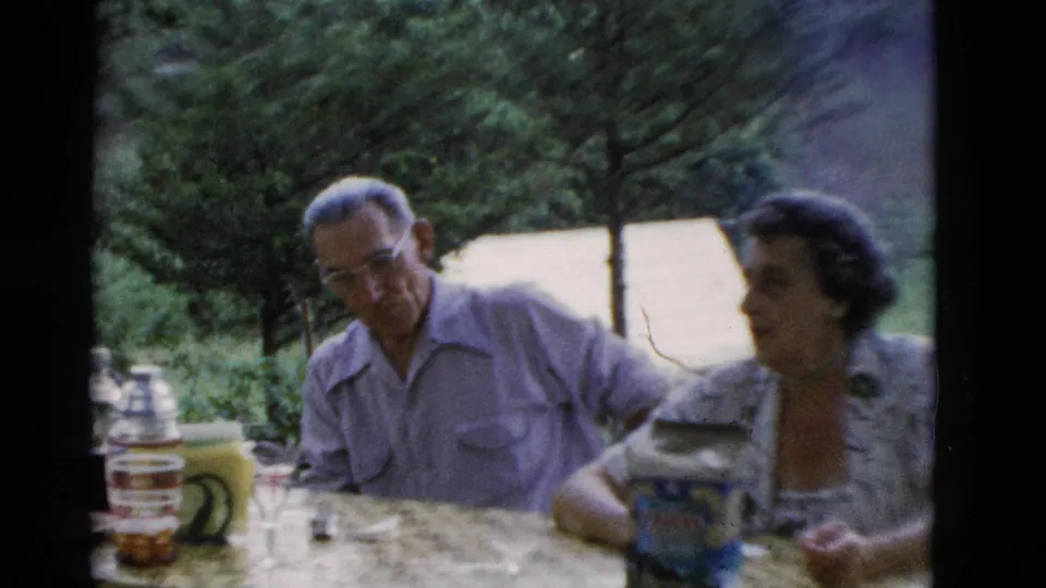 A man and woman sit at a picnic table in 1975 Somerset, Kentucky footage, with cups, a jug, and packaged food visible.