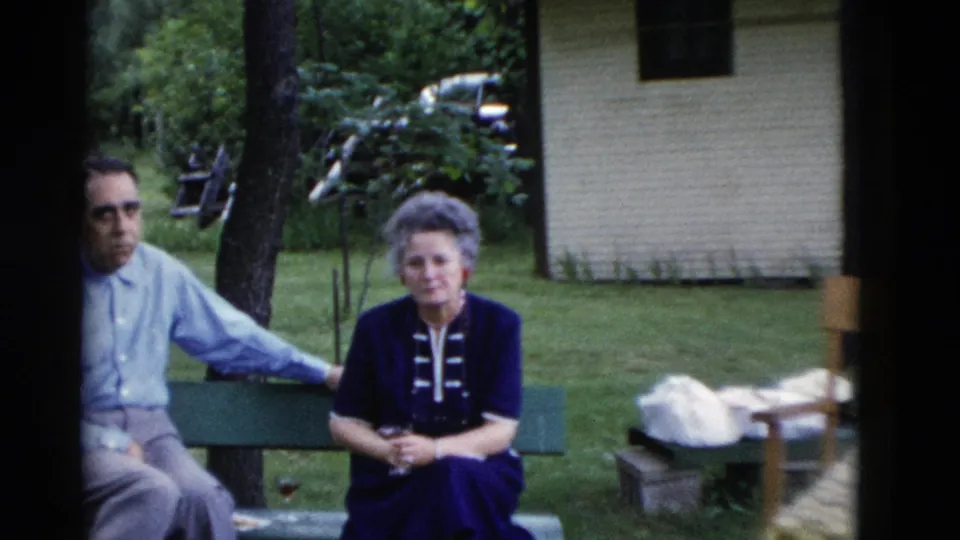A man and woman sit on a green bench at the Somerset reunion, with a small light-colored building and parked car in the background.