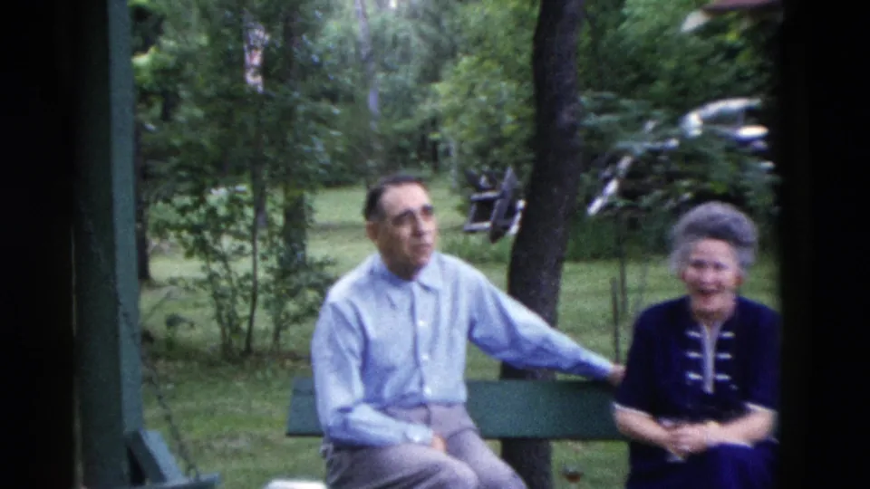 A man and woman seated on a green bench face the camera in soft 8mm footage from the supplied 1975 Somerset, Kentucky setting.