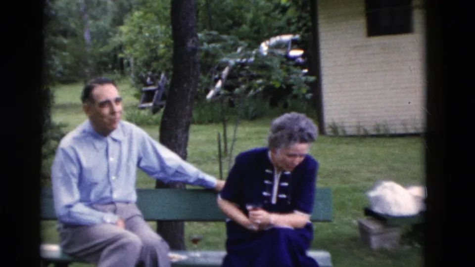 The man remains seated with one arm along the bench while the woman looks down, with trees and a light building visible behind them.