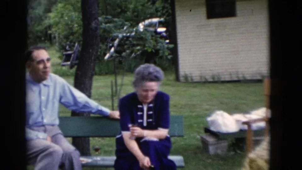 In 1975 Somerset, Kentucky home-movie footage, an older man and woman sit together on a green bench in a wooded yard.