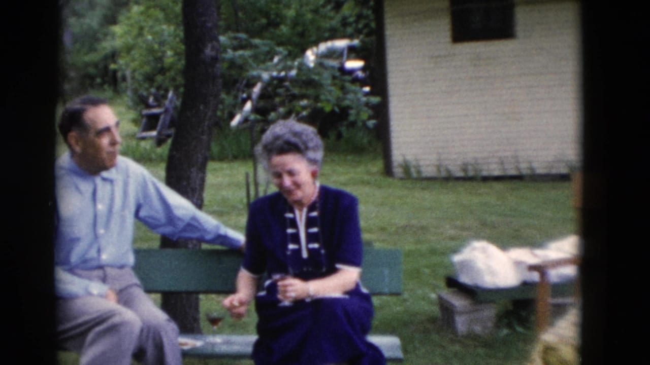 The older couple remains seated on the bench, with the woman holding a glass and the man turned toward her in the yard.