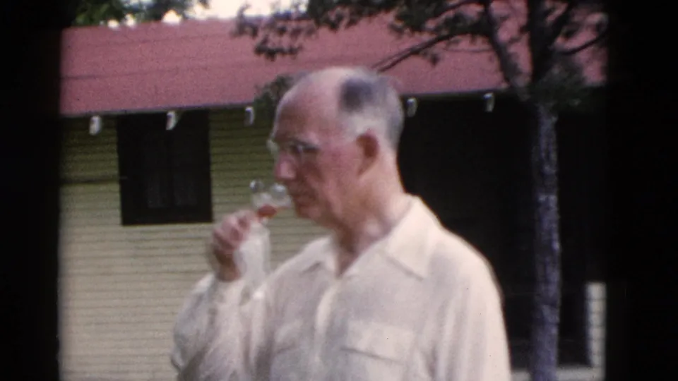 An older man wearing glasses drinks from a glass in the yard, with a small building and red roof behind him.