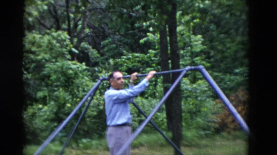 The older man stands by a metal exercise or play frame in the wooded yard, holding the bar with both hands.
