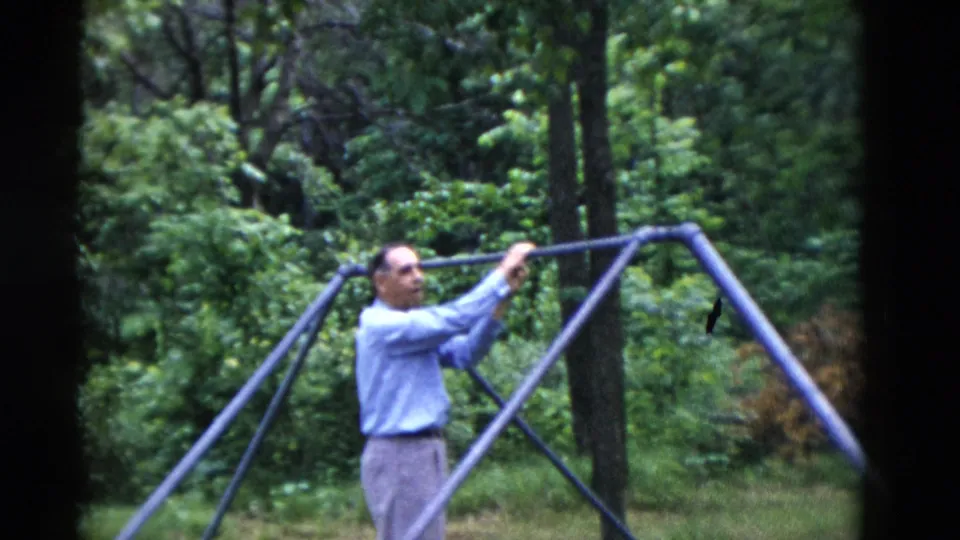 In 1975 Somerset, Kentucky, an older man in a blue dress shirt and gray trousers reaches toward a purple metal swing-set bar in a green backyard.