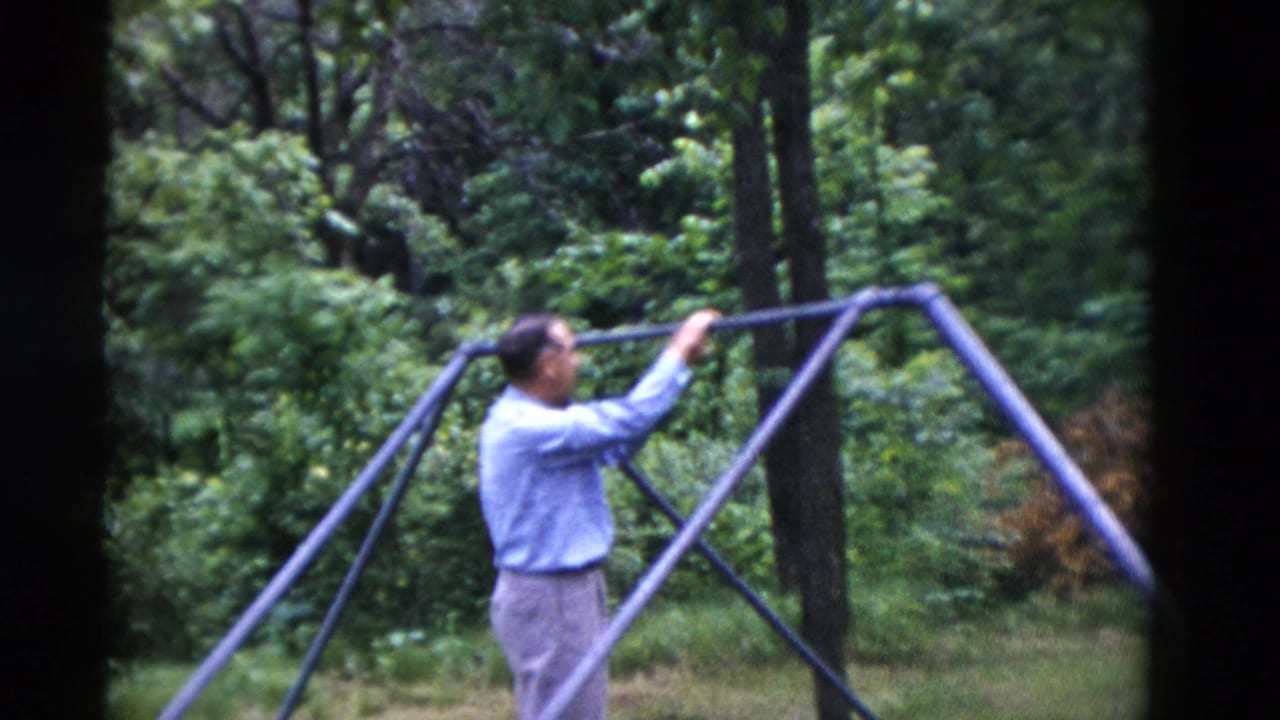 In the Somerset, Kentucky home-movie frame, the man reaches up to hold the purple swing-set bar with trees and grass behind him.