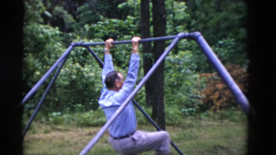 The 1975 8mm frame shows the man hanging from the swing-set bar, with his blue shirt and gray trousers visible against dense green foliage.