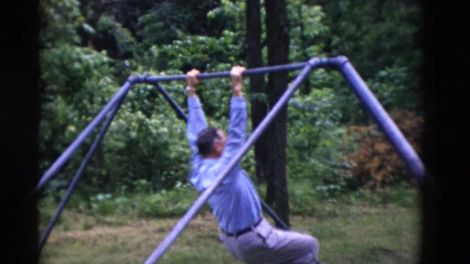 In Somerset, Kentucky 1975 footage, the man lifts himself on the purple metal swing-set bar, framed by trees and lawn.