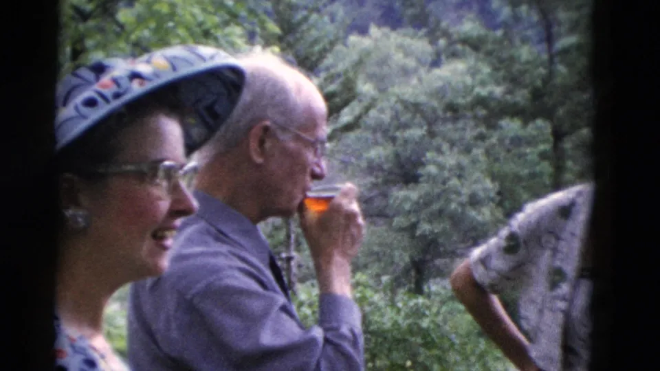 A woman in glasses smiles as an older man drinks from a small glass in wooded 1975 Somerset home-movie footage.
