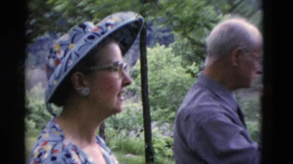 A woman in a decorated hat and glasses stands beside an older man in profile in wooded Somerset, Kentucky footage from 1975.