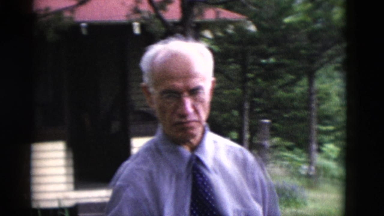 Older man in a light shirt and dark tie standing outdoors near trees and a house.