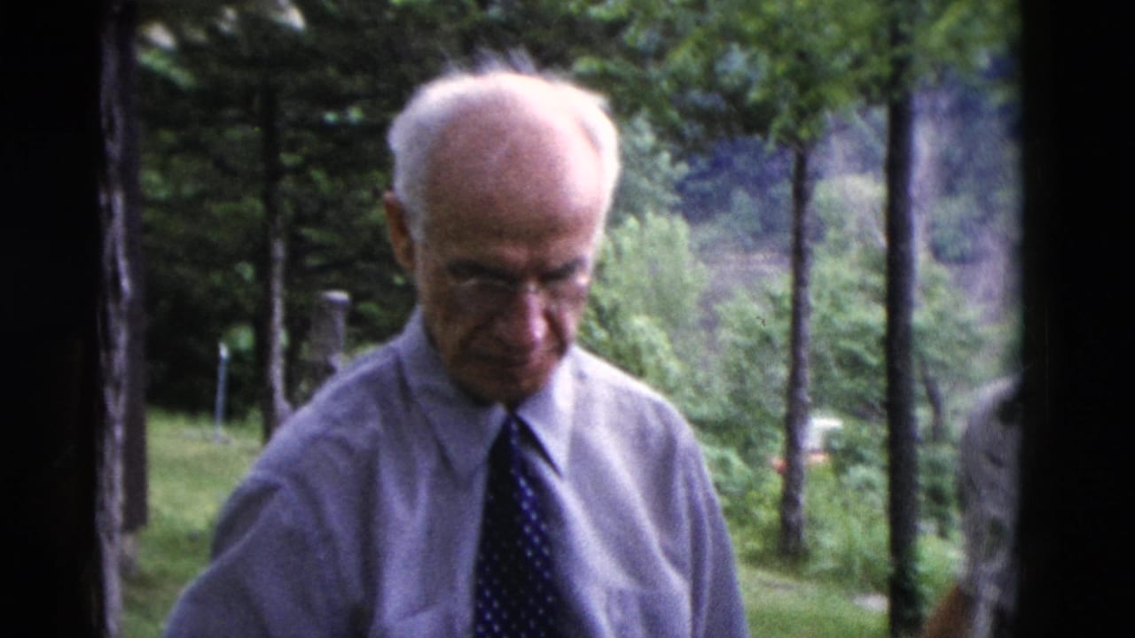 Older man in a shirt and tie looks downward in the green yard during the 1975 Somerset home-movie scene.
