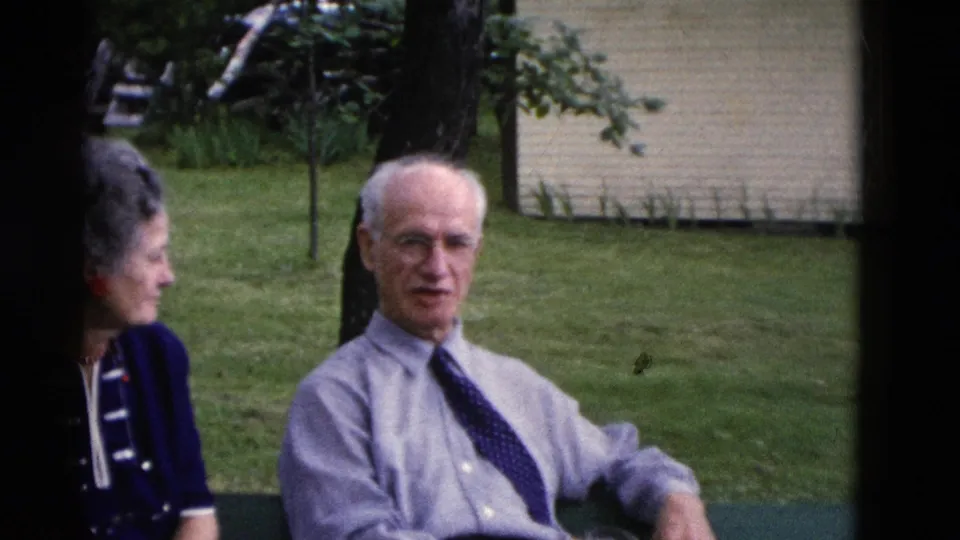 An older man in a light shirt and patterned tie sits beside a woman in a blue dress during a 1975 Somerset backyard gathering.
