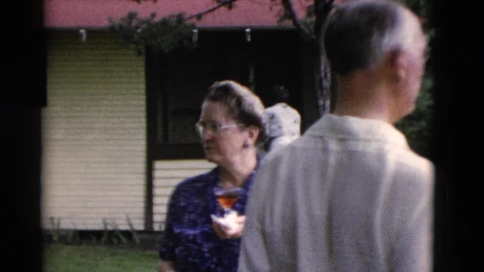 A woman holding a small cup stands in a grassy Somerset yard while an older man is seen from behind in 1975 home-movie footage.