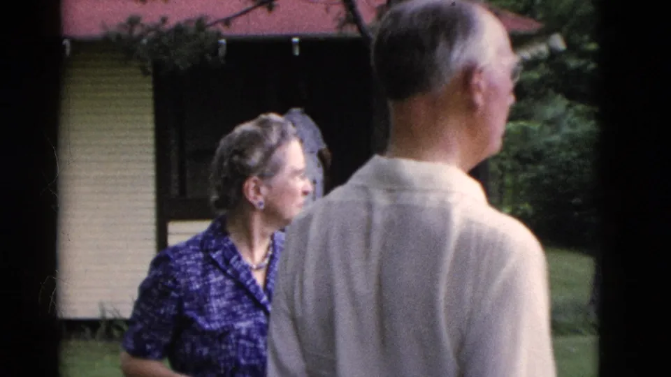 A woman in a blue patterned dress and an older man seen from behind look away across a Somerset, Kentucky backyard in 1975 footage.