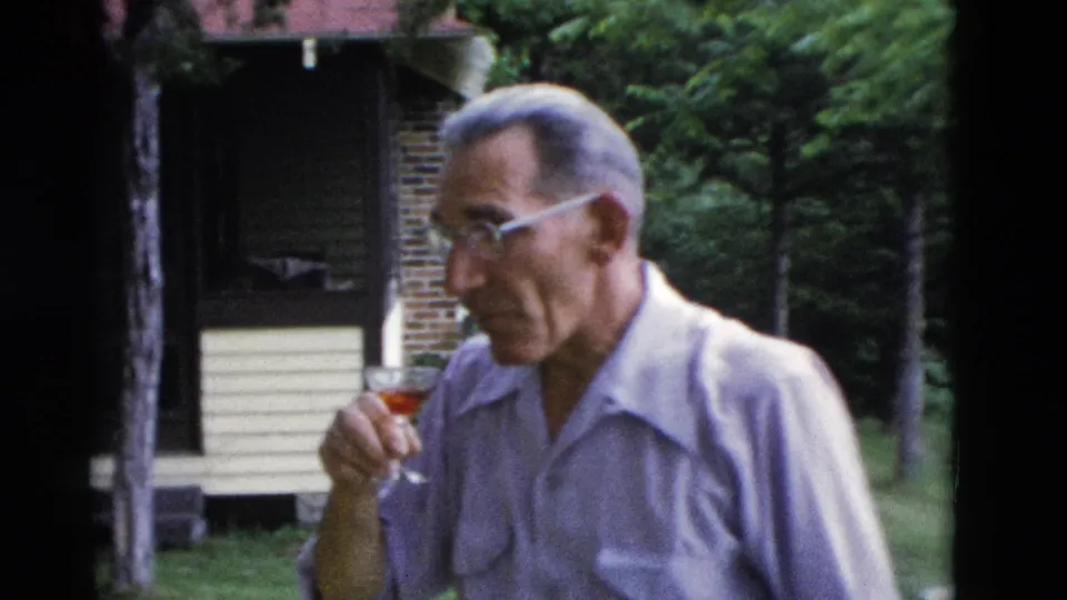 An older man in glasses raises a small stemmed drink on a lawn beside a yellow-sided house in 1975 Somerset, Kentucky home-movie footage.